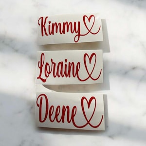 May include: Three white rectangular stickers with red cursive text and a heart symbol. The stickers read "Kimmy", "Loraine", and "Deene". The stickers are arranged vertically on a white surface.