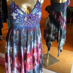 May include: Two dresses on mannequins. The first dress has a blue lace bodice and a tie-dye skirt with pink, blue, and purple hues. The second dress has a dark blue bodice and a tie-dye skirt with purple and gray tones.
