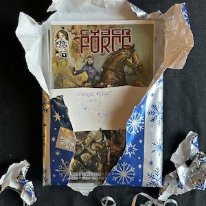 May include: A comic book titled "Cyber Force" is partially visible, wrapped in blue and white snowflake-patterned paper. The comic features a woman on a horse. The wrapping is torn, revealing the comic and handwritten text.