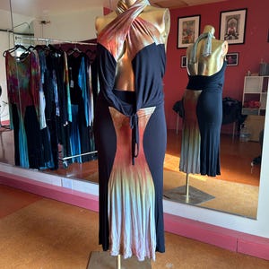 May include: A black maxi dress with a halter neckline and a tie-waist detail. The dress features a color-blocked design with a tie-dye pattern in shades of orange, red, and yellow. The dress is displayed on a gold mannequin.