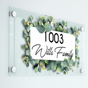 May include: A clear glass door sign with a white background and green eucalyptus leaves surrounding the text "1003 Wills Family".