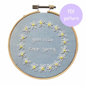 Puede incluir: Un aro de bordado azul con un diseño de bordado blanco de una corona de margaritas con el texto "You nice Keep going" en el centro.