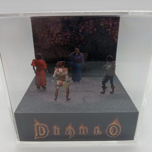 May include: A clear acrylic display case featuring four pixelated figures from the Diablo video game. The figures stand on a gray platform with the Diablo logo at the base. The background shows a blurred forest scene.