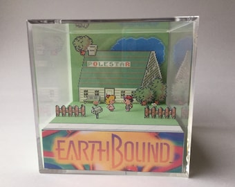May include: A diorama of a green house with a sign that reads "POLESTAR" and two pixelated figures standing in front of it. The diorama is inside a clear acrylic cube and has a base with the words "EARTHBOUND" printed on it.