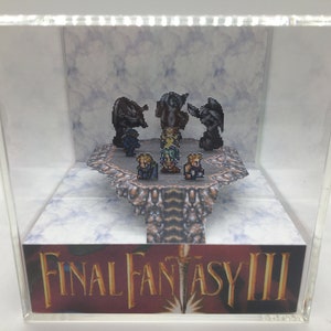 May include: A clear acrylic display case with a 3D diorama of characters from the video game Final Fantasy III. The diorama features a group of pixelated characters standing on a platform with a background of clouds. The base of the display case features the text "FINAL FANTASY III".