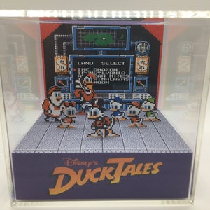 May include: A pixelated diorama featuring characters from Disney's DuckTales. The scene includes Scrooge McDuck and his nephews in a retro video game style, set against a backdrop with the words "Land Select". The base of the display reads "Disney's DuckTales".