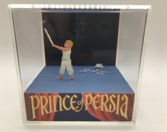 May include: A 3D diorama of a scene from the classic video game Prince of Persia. The diorama features a pixelated character holding a sword, a skull, and a blue background. The diorama is encased in a clear acrylic box.