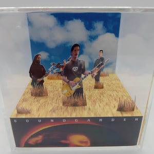 May include: A clear acrylic display featuring a band scene with three figures holding guitars on a wheat field stage. The background shows a blue sky with clouds. The band's name, SOUNDGARDEN, is printed at the bottom.