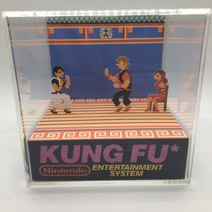 May include: A clear acrylic display case with a 3D diorama of the classic Nintendo Entertainment System game Kung Fu. The diorama features pixelated characters in a fighting pose on a wooden floor with a blue background.