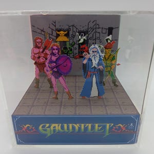 May include: A 3D diorama of the classic arcade game Gauntlet. The diorama features five pixelated characters, including a warrior, a wizard, a valkyrie, an elf, and a sorceress, standing on a stone floor. The background is a gray stone wall with a dungeon-like design. The diorama is in a clear plastic case with a blue base that reads "GAUNTLET EX".