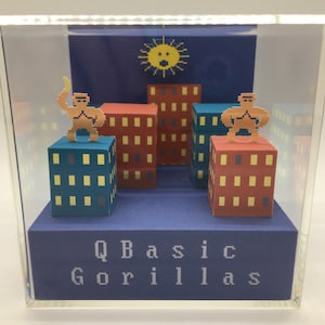 May include: A diorama of a pixelated city scene with two figures standing on top of buildings. The figures are brown and yellow and the buildings are red, blue, and yellow. The diorama is in a clear acrylic box with a blue base that reads "QBasic Gorillas".