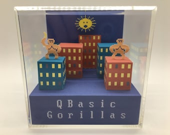 May include: A diorama of a pixelated city scene with two figures standing on top of buildings. The figures are brown and yellow and the buildings are red, blue, and yellow. The diorama is in a clear acrylic box with a blue base that reads "QBasic Gorillas".