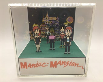 May include: A 3D diorama of the classic video game "Maniac Mansion". The diorama features the main characters from the game, including a woman in a black dress, a man in a pink shirt, and a man in a blue shirt. The diorama is set against a backdrop of the mansion from the game, with a full moon in the sky.