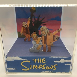 May include: A diorama featuring characters from "The Simpsons" in front of a Statue of Liberty silhouette. The scene is enclosed in a clear acrylic box. The base is blue with white clouds and the words "THE SIMPSONS" in yellow.