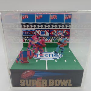 May include: A miniature diorama of a football game with pixelated figures in red, blue, and white uniforms. The diorama is set in a stadium with a green field, white lines, and a blue and white 'TECHNO' logo. The background features a blue and white pixelated pattern with American flags. The diorama is enclosed in a clear acrylic case and has the text 'SUPER BOWL' on the front.