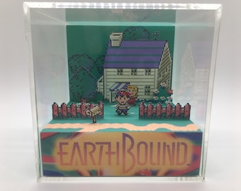 May include: A pixelated diorama of a scene from EarthBound, featuring a house with a purple roof, a picket fence, and two characters. The diorama is encased in a clear acrylic box. The EarthBound logo is visible at the bottom.