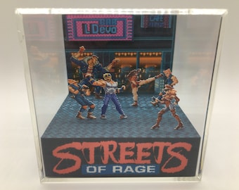 Puede incluir: Un diorama 3D del clásico juego de arcade Streets of Rage. El diorama presenta personajes pixelados del juego, incluyendo un personaje femenino con un atuendo rojo y negro, un personaje masculino con una camisa azul y jeans, y un personaje masculino con una camisa negra y jeans. El diorama está ambientado en el fondo de una calle de la ciudad con un bar y una cafetería.
