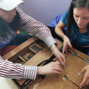 Puede incluir: Dos personas trabajan juntas para ensamblar una caja de madera con una tapa con bisagras. La caja tiene un peque&ntilde;o compartimento con una cerradura y un cable negro conectado al interior. La persona de la izquierda lleva una gorra de b&eacute;isbol blanca y una camisa a rayas. La persona de la derecha lleva una camisa azul y tiene el pelo largo y oscuro.