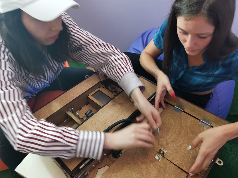 Puede incluir: Dos personas trabajan juntas para ensamblar una caja de madera con una tapa con bisagras. La caja tiene un peque&ntilde;o compartimento con una cerradura y un cable negro conectado al interior. La persona de la izquierda lleva una gorra de b&eacute;isbol blanca y una camisa a rayas. La persona de la derecha lleva una camisa azul y tiene el pelo largo y oscuro.