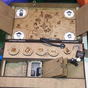 Puede incluir: Un juego de cofre del tesoro de madera con tem&aacute;tica pirata. La parte superior del cofre tiene un mapa con una calavera y huesos cruzados y una br&uacute;jula. La parte inferior del cofre tiene cinco piezas de rompecabezas redondas de madera y una cerradura con una llave.