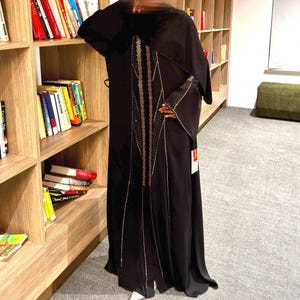 May include: A black abaya with gold and bronze embellishments. The long-sleeved garment features intricate detailing down the front and on the sleeves. The abaya is floor-length and appears to be made of a flowing fabric.