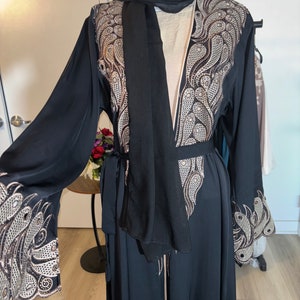 May include: A black abaya with a matching black scarf. The abaya has intricate silver and gold embroidery on the sleeves and neckline.