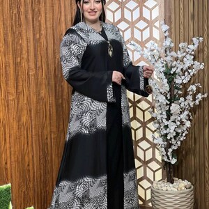 May include: A black abaya with a white geometric pattern. The abaya has long sleeves and a long, flowing skirt. The abaya is worn by a person standing in front of a wooden wall.