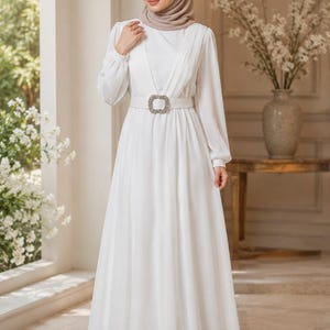 May include: A floor-length white dress with long sleeves, featuring vertical pleats on the bodice and a wide matching belt with a large, rectangular, crystal-embellished buckle at the waist. The dress has a high neckline and a flowing skirt.