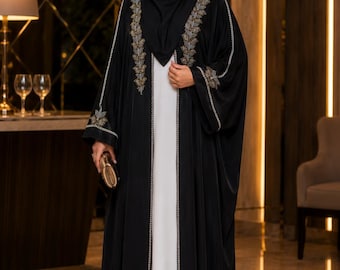 Könnte beinhalten: Eine schwarze, vorne offene Abaya mit aufwendigen gold- und silberfarbenen Blattstickereien an den Revers und Ärmelbündchen, getragen über einem bodenlangen weißen Kleid. Das Outfit umfasst ein passendes schwarzes Kopftuch und eine goldene Clutch.
