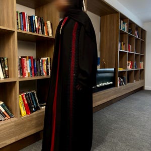 May include: A black abaya with red and sequin accents. The abaya features a vertical red stripe down the front and is adorned with sequins. The garment is long and flowing, suitable for modest wear.