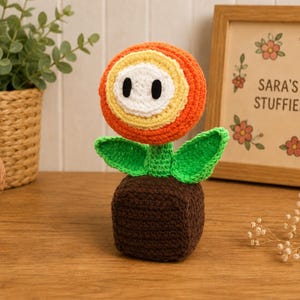 May include: A crocheted fire flower toy featuring a brown square base, a green stem with two leaves, and a circular head with orange, yellow, and white concentric rings and two black oval eyes. The yarn texture is visible throughout the entire object.