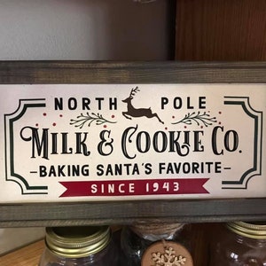 May include: A rustic wooden sign with a white background and black lettering. The sign reads "North Pole Milk & Cookie Co. - Baking Santa's Favorite - Since 1943". A red ribbon is below the text. A black silhouette of a reindeer is above the text.
