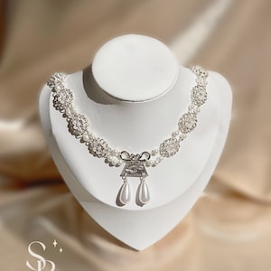 May include: A pearl necklace with floral accents and a silver pendant with two teardrop-shaped pearl drops. The necklace is displayed on a white mannequin against a soft, neutral background. The necklace is a delicate piece of jewelry.