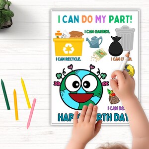 A child's hand coloring the Earth Day activity sheet, which is now shown in color with blue and green Earth, yellow recycling bin, and colorful text. Colored pencils are visible in the corner with a potted plant in the background on a white wooden surface.