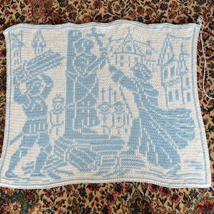 May include: A knitted throw blanket featuring a blue and white design of a building and a person. The blanket is crafted from a soft yarn and has a textured surface.