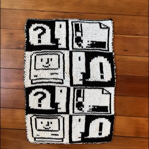May include: A black and white knitted blanket with a pixelated design of computer icons. The icons include a floppy disk, a question mark, and a computer monitor.