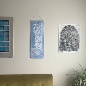 以下が含まれることがあります： 中央に人物が描かれた青と白のニットの壁掛け。壁掛けは、白い壁に額縁に入った写真と、古い碑文の額縁に入ったプリントが飾られています。