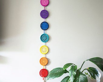 May include: A colorful, handmade crochet wall hanging featuring seven circular designs in a rainbow of colors: pink, purple, blue, turquoise, yellow, orange, and red. The wall hanging is suspended from a loop at the top and includes a tassel at the bottom. The decor is displayed on a light gray wall.