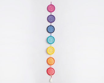 May include: A colorful chakra wall hanging featuring seven circular, crocheted medallions in a rainbow of colors: pink, purple, blue, turquoise, yellow, orange, and red. Each medallion has a unique design and is connected by a string. A white tassel hangs at the bottom.