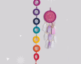 May include: Two dream catchers hanging on a wall. One is a vertical string of seven colorful rings, and the other is a pink crocheted circle with feathers and beads. The rings are in rainbow colors.