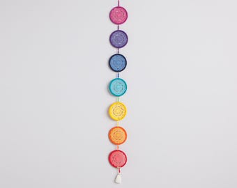 May include: A wall hanging with seven colorful crocheted circles, each representing a chakra, hanging in a vertical line. The circles are red, orange, yellow, green, blue, indigo, and violet.