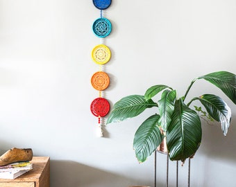 May include: A colorful, handmade wall hanging featuring seven crocheted circles in a rainbow of colors: pink, purple, blue, turquoise, yellow, orange, and red. The circles are connected by a chain and hang vertically. A white tassel hangs from the bottom.