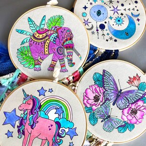 May include: Three embroidery hoops with colorful designs. The first hoop features a purple elephant with intricate patterns, the second hoop has a blue and white moon and stars design, and the third hoop has a purple butterfly and flowers.
