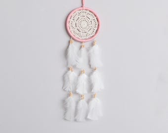 May include: A dream catcher featuring a circular pink hoop wrapped in thread with a white crocheted doily center. Three strands of white feathers hang from the bottom, each accented with small light brown wooden beads at the attachment points.