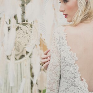 May include: A woman wearing a white lace dress with a low back and a silver headband. She is standing in front of a wall of dreamcatchers with white feathers.