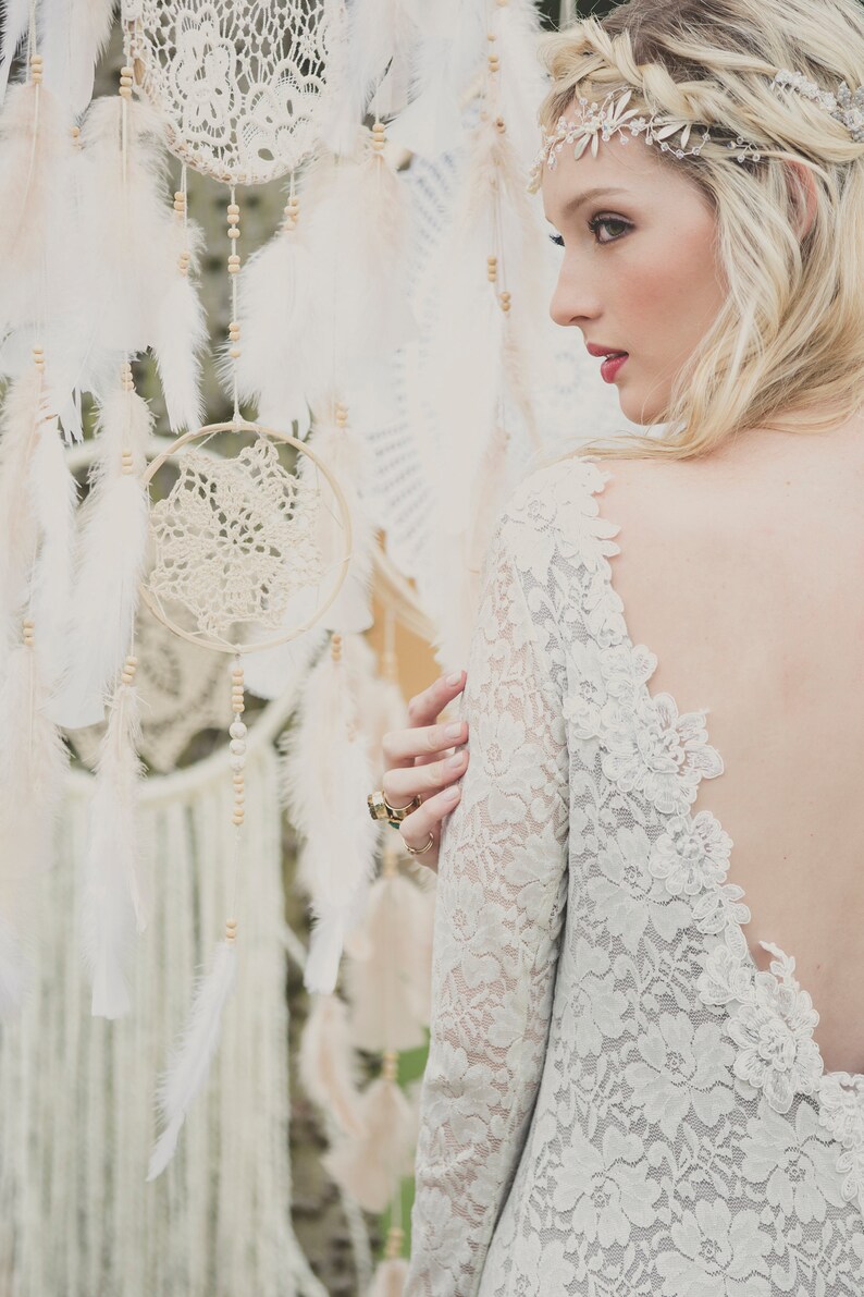 May include: A woman wearing a white lace dress with a low back and a silver headband. She is standing in front of a wall of dreamcatchers with white feathers.