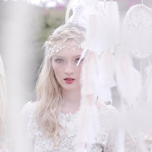 May include: A woman with blonde hair wearing a white lace dress and a silver headband with rhinestones. She is standing in front of a white background with white dreamcatchers hanging from the ceiling.