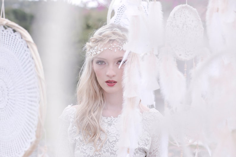 May include: A woman with blonde hair wearing a white lace dress and a silver headband with rhinestones. She is standing in front of a white background with white dreamcatchers hanging from the ceiling.