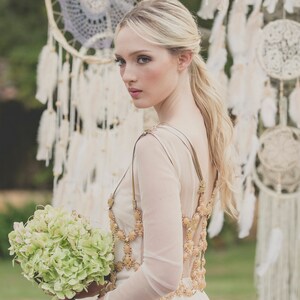 May include: A bride wearing a white dress with a gold beaded bodice and a bouquet of green hydrangeas. She is standing in front of a backdrop of dreamcatchers.