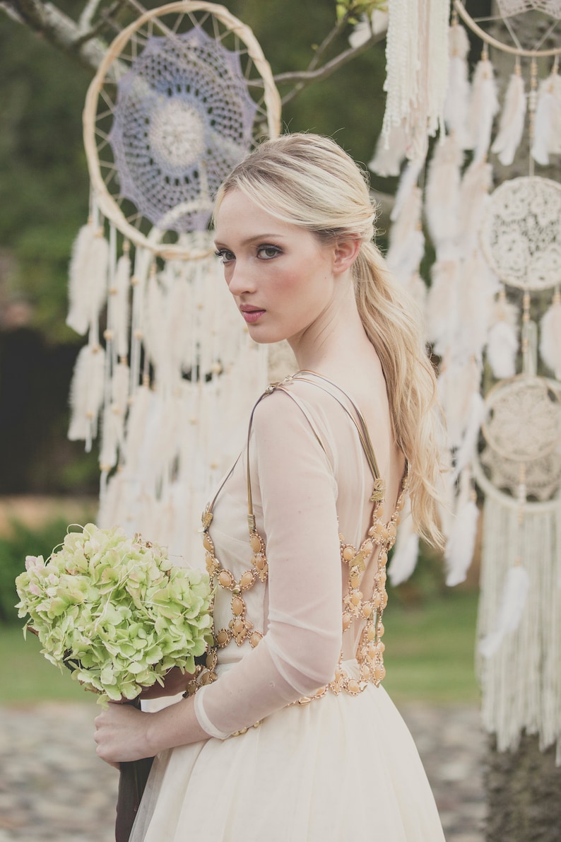 May include: A bride wearing a white dress with a gold beaded bodice and a bouquet of green hydrangeas. She is standing in front of a backdrop of dreamcatchers.